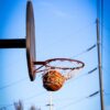 A basketball perfectly swishes through the hoop on an outdoor court against a clear blue sky.