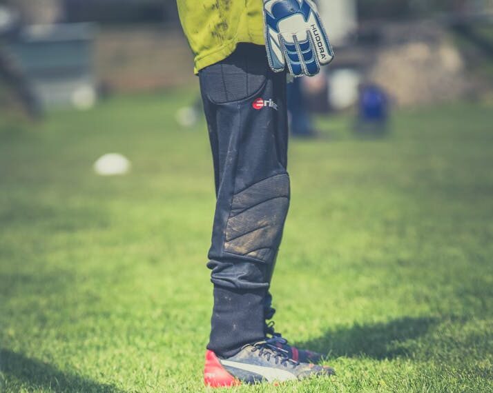 Goalkeeper standing on green field, focused on legs with gear, during a sunny day soccer match.