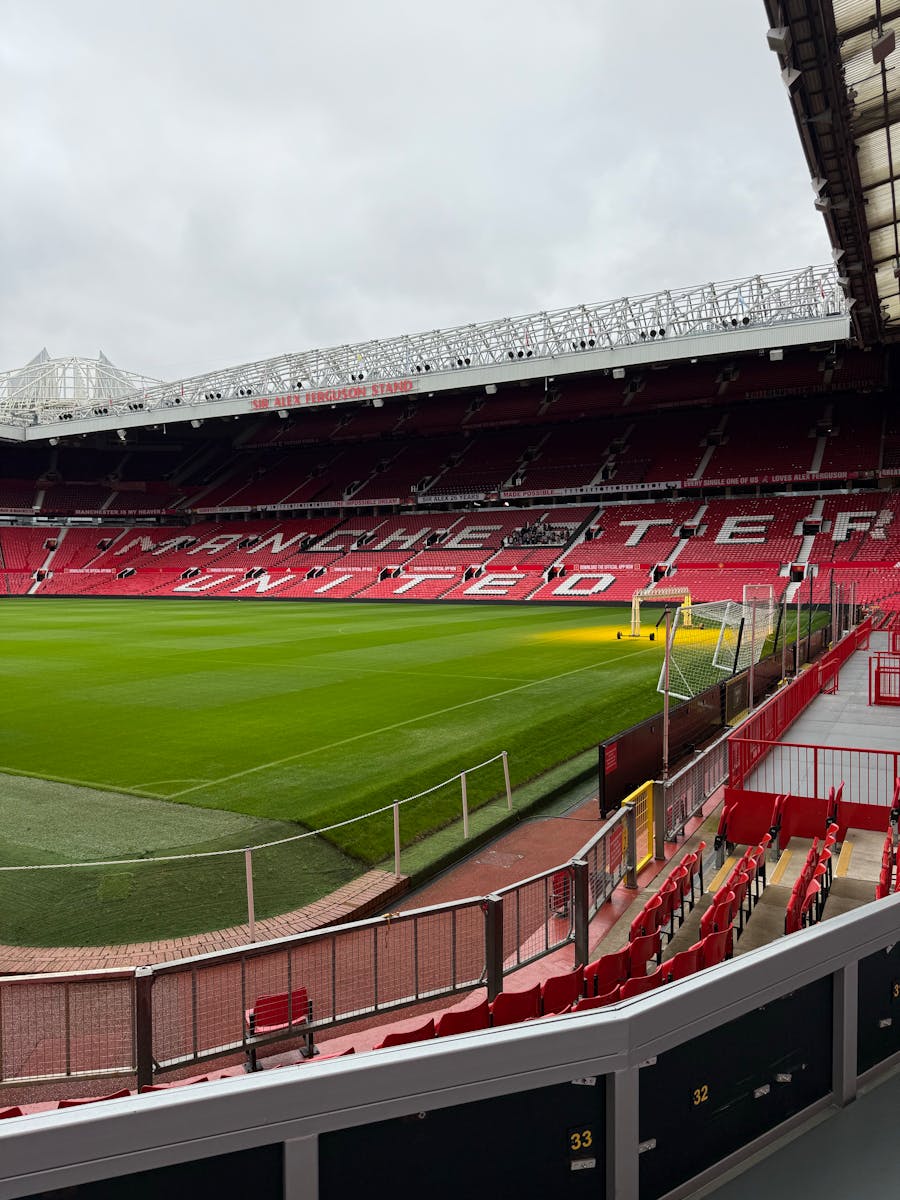 Empty Old Trafford stadium with red seats and green field under cloudy skies.