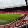 Empty Old Trafford stadium with red seats and green field under cloudy skies.