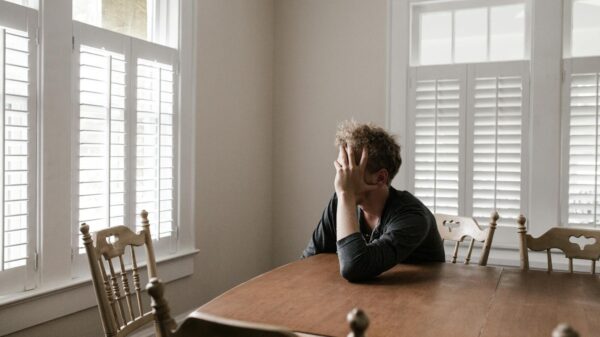 A man sits alone at a table in a bright room, displaying deep contemplation.