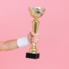 A hand holding a golden trophy against a vibrant pink background, symbolizing victory and success.