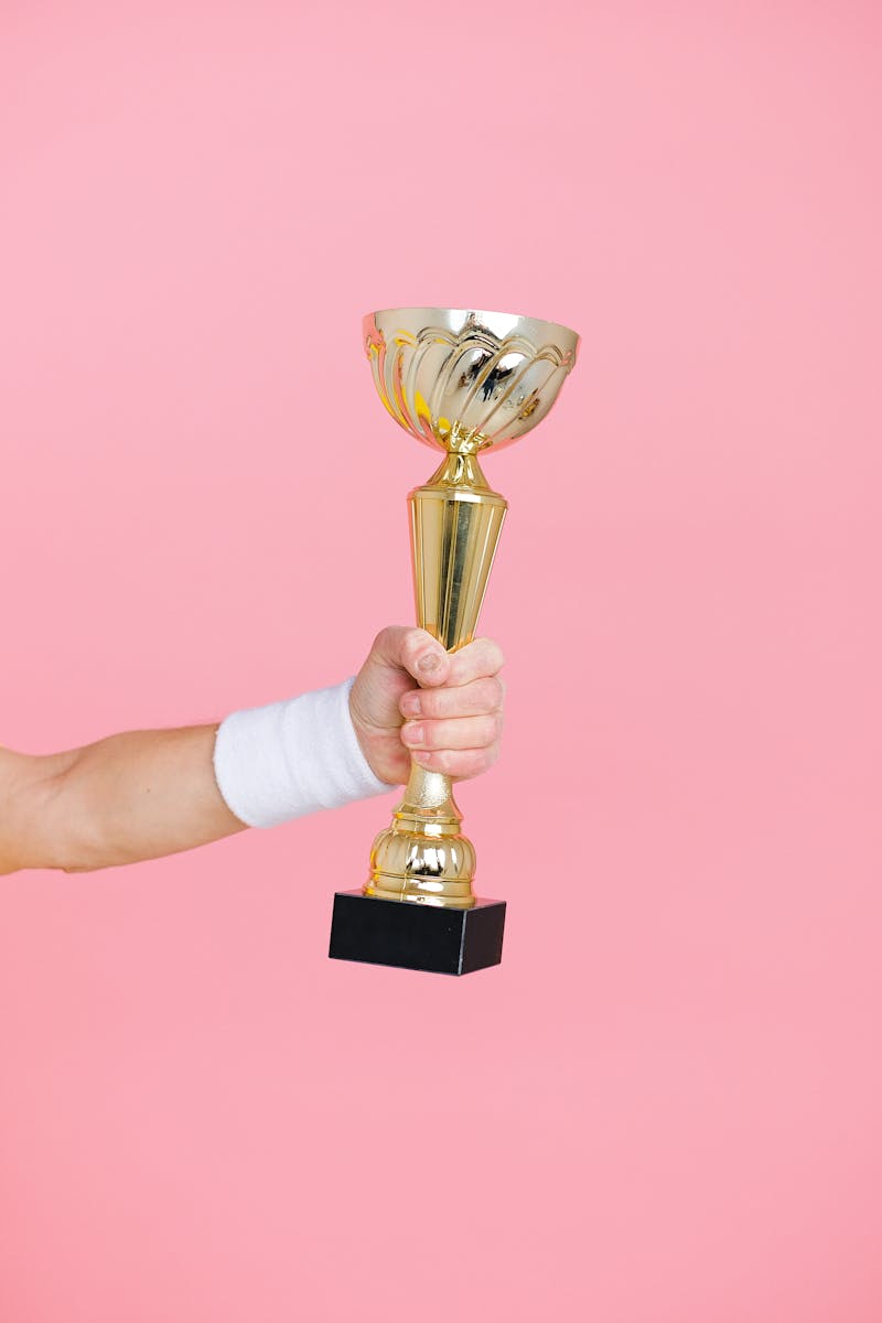 A hand holding a golden trophy against a vibrant pink background, symbolizing victory and success.
