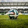 A vibrant soccer ball rests on a lush green field inside an empty stadium, capturing the essence of sports.