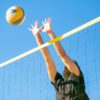 Dynamic outdoor volleyball game with a male player blocking a ball under clear blue skies.