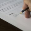 A hand signs a formal contract with a pen on a wooden desk.