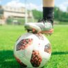 Close-up of a soccer ball with a player's foot on it, set on a vibrant green soccer field during a sunny day.