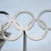 The iconic Olympic rings in front of a tall structure on a clear day.