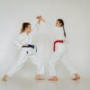 Two women engage in martial arts training, wearing traditional karate uniforms indoors.