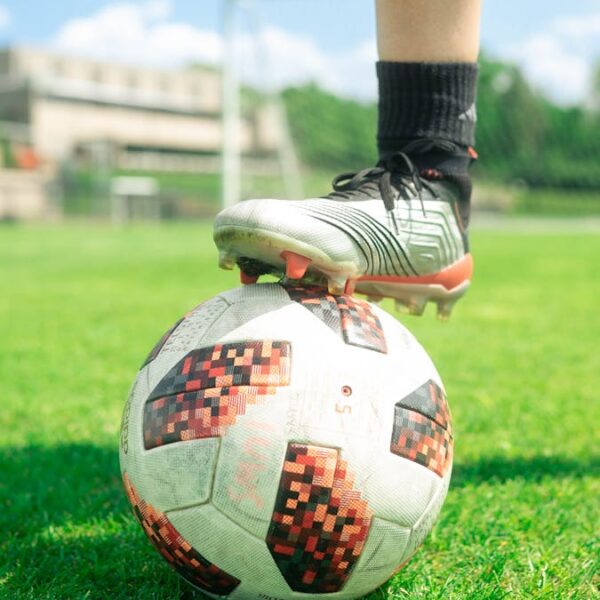 Close-up of a soccer ball with a player's foot on it, set on a vibrant green soccer field during a sunny day.