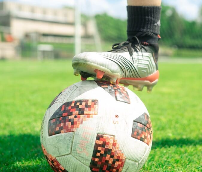 Close-up of a soccer ball with a player's foot on it, set on a vibrant green soccer field during a sunny day.