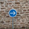 A blue directional sign with an arrow against a rustic stone wall in an urban setting.