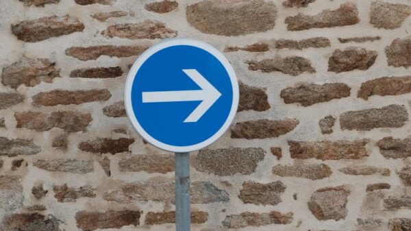 A blue directional sign with an arrow against a rustic stone wall in an urban setting.