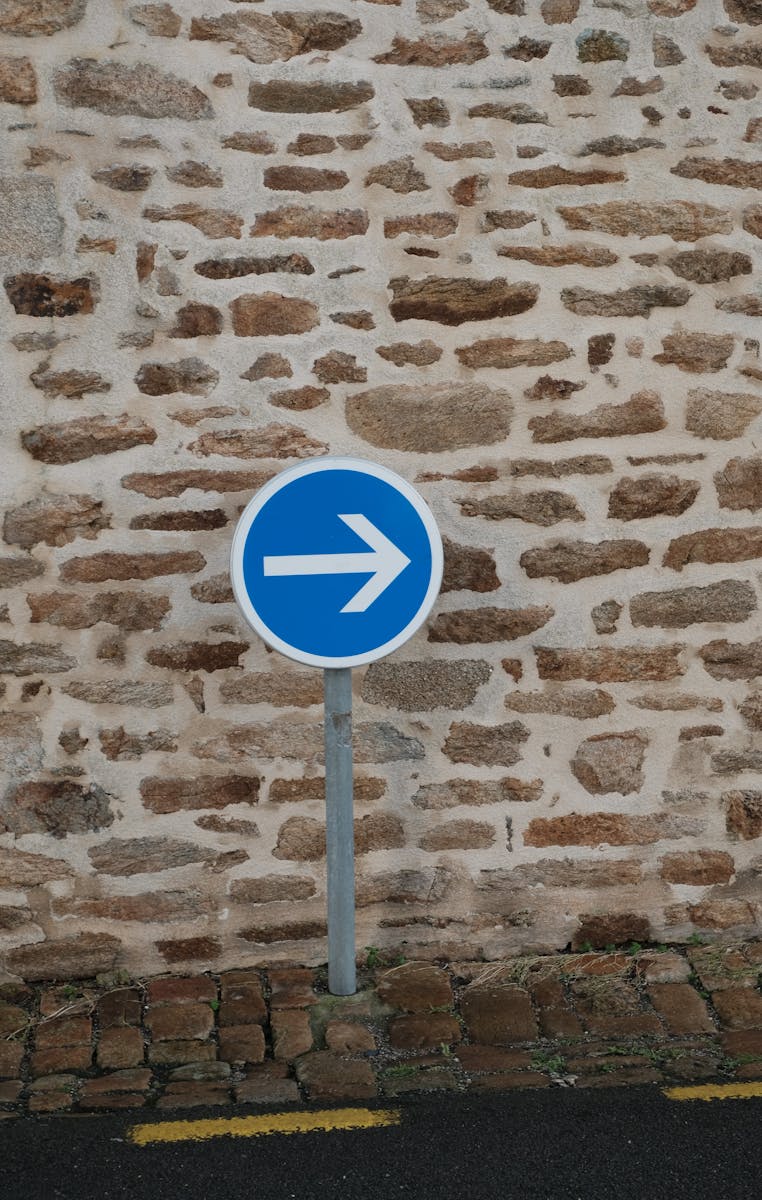 A blue directional sign with an arrow against a rustic stone wall in an urban setting.