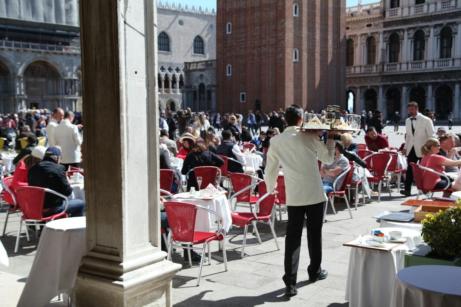 Busy outdoor cafe in Piazza San Marco, Venice with waiters in white jackets serving guests.
