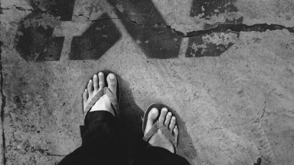 Black and white of crop unrecognizable person in dark outfit and flip flops standing on asphalt road with graffiti