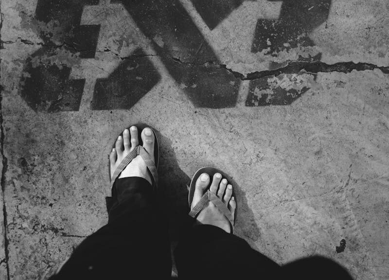 Black and white of crop unrecognizable person in dark outfit and flip flops standing on asphalt road with graffiti