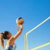 A woman plays volleyball on a sunny day, reaching to hit the ball over the net.