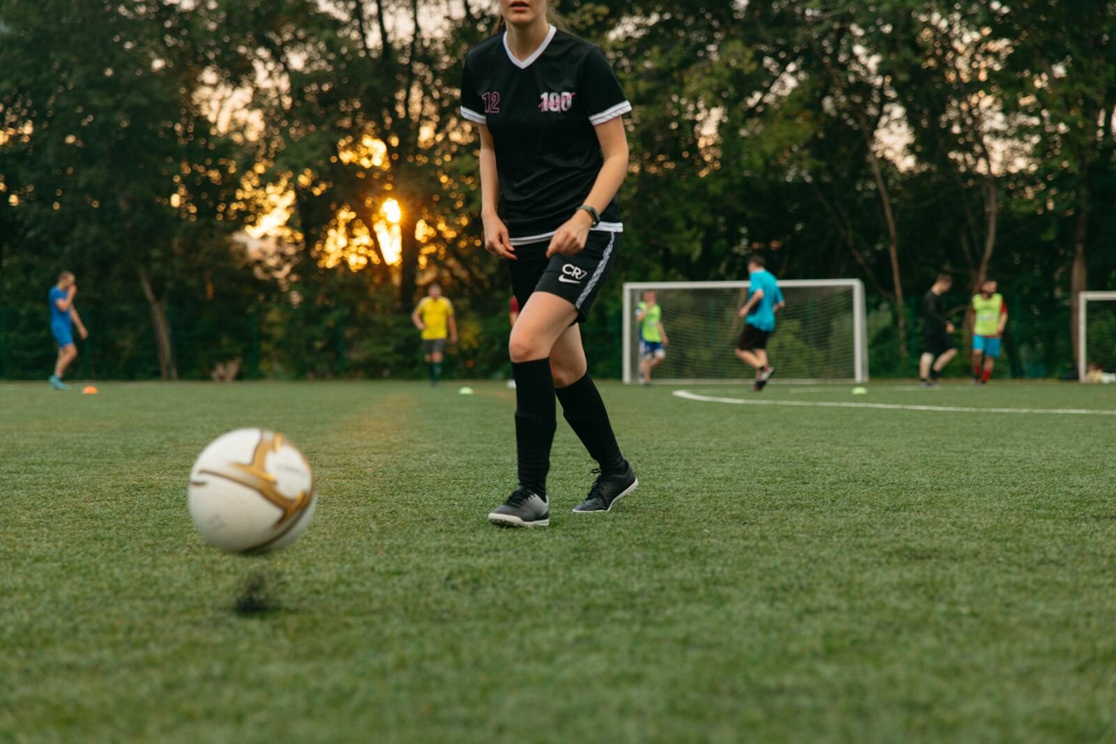 Female soccer player dribbling a ball during a casual game on a grassy field at sunset.