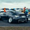 A sleek black sports car parked at an outdoor auto event with spectators and other vehicles in the background.
