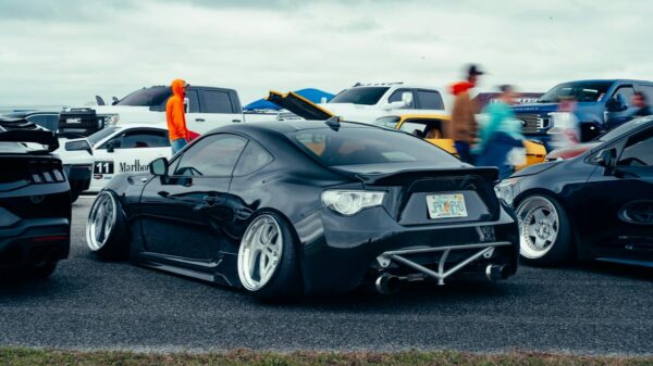 A sleek black sports car parked at an outdoor auto event with spectators and other vehicles in the background.