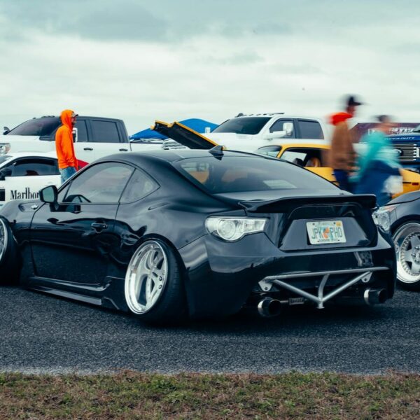 A sleek black sports car parked at an outdoor auto event with spectators and other vehicles in the background.