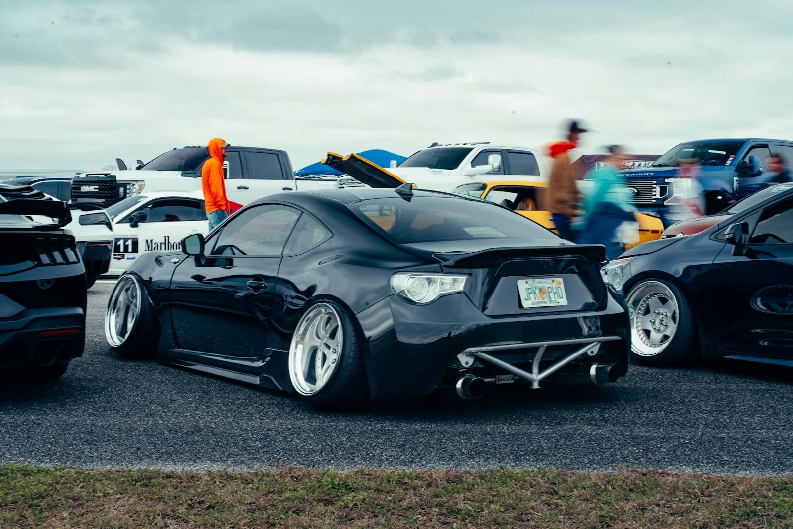 A sleek black sports car parked at an outdoor auto event with spectators and other vehicles in the background.