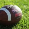 Detailed image of a Wilson NCAA football resting on vibrant green grass in daylight.