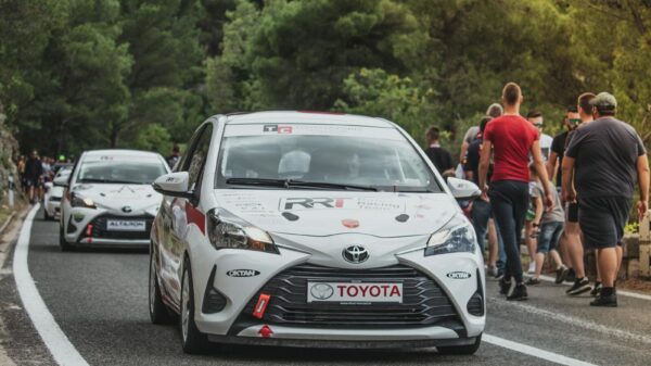 Toyota Yaris rally car leads a motorsport event on a scenic forest road with spectators.