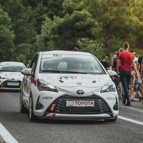 Toyota Yaris rally car leads a motorsport event on a scenic forest road with spectators.