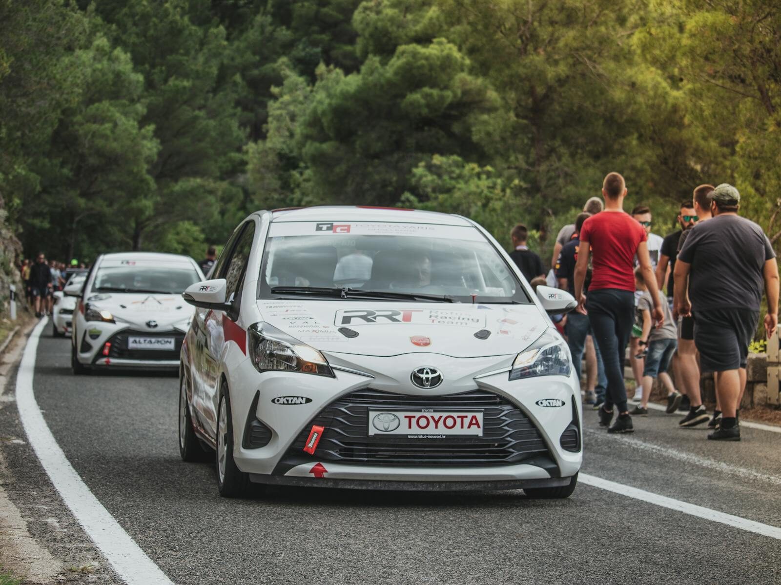 Toyota Yaris rally car leads a motorsport event on a scenic forest road with spectators.