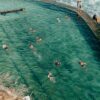 High angle of unrecognizable tourists in swimsuit swimming in pool near pier with railing and waving ocean with rocky formations in sunny summer day