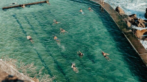 High angle of unrecognizable tourists in swimsuit swimming in pool near pier with railing and waving ocean with rocky formations in sunny summer day