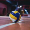 Yellow and white volleyball resting on an indoor volleyball court with bright lights.