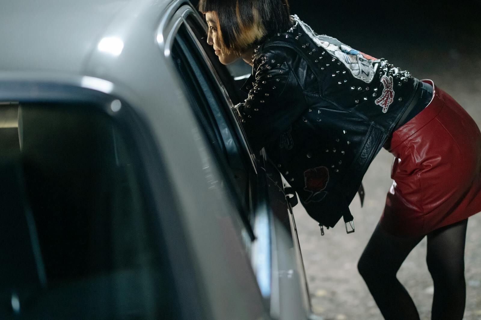 Side view of a woman in a leather jacket leaning on a car at night.