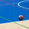 A basketball rests on a vibrant blue indoor court with wood flooring and colorful lines.