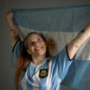 Smiling woman holding up the Argentina flag in celebration, wearing a blue and white jersey.