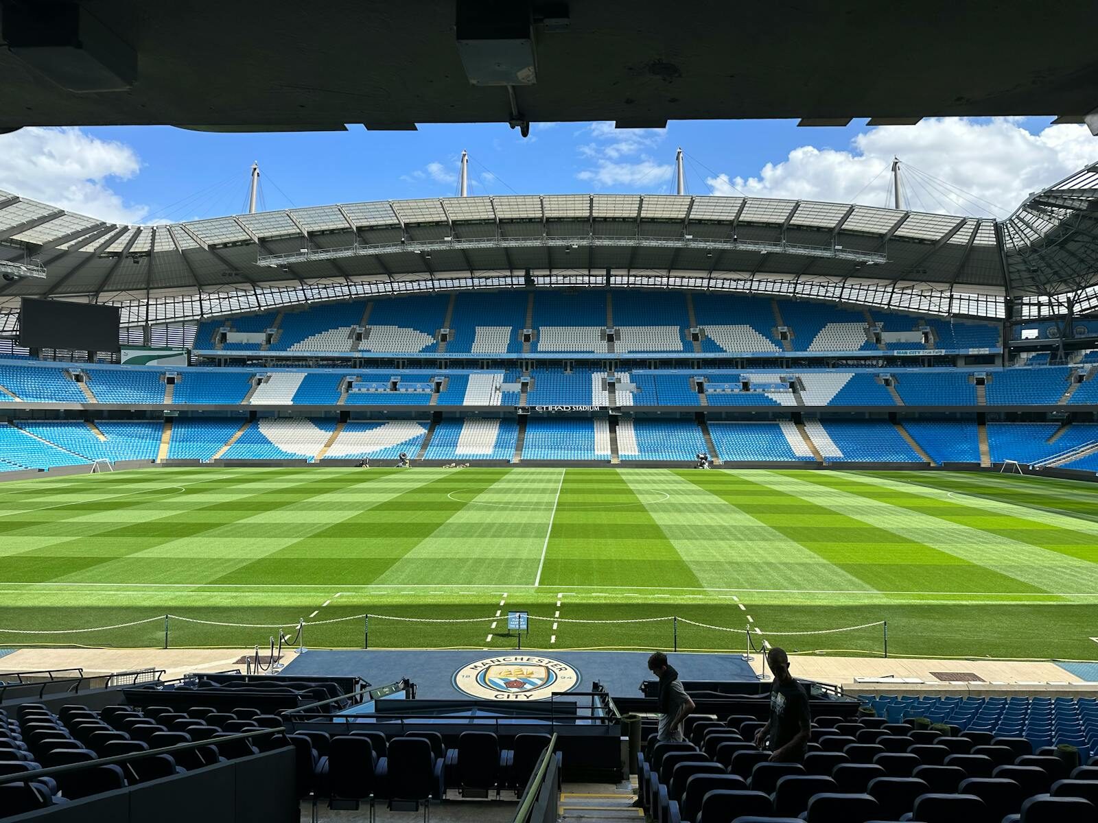 Panoramic view of Etihad Stadium, showcasing the empty football pitch and stands under a clear blue sky.