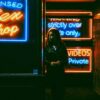 woman in black jacket standing in front of blue and red UNKs neon signage