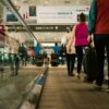 Passengers walking on a moving walkway in a busy airport terminal with signs for gates.