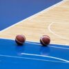 Two basketballs resting on a blue indoor basketball court with wooden flooring.