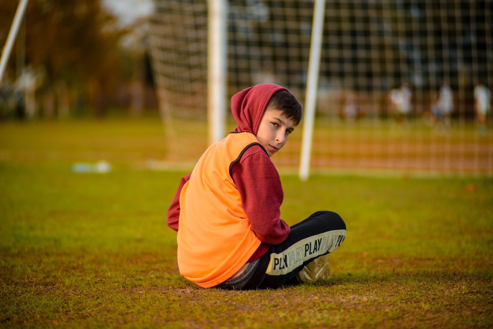 Boy in hoodie seated on a football field in Antalya, gazing over shoulder.